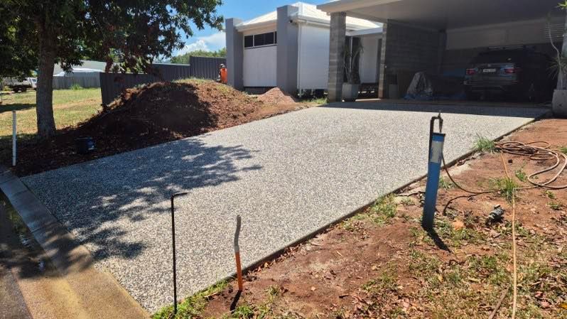 Fresh concrete driveway leading to a covered carport beside a white house, with red dirt landscaping. — King Reef Concrete in Kurrimine Beach, QLD