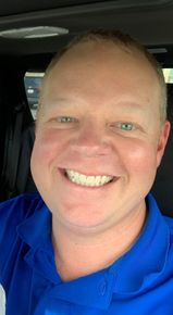 A man in a blue shirt is smiling while sitting in a car.