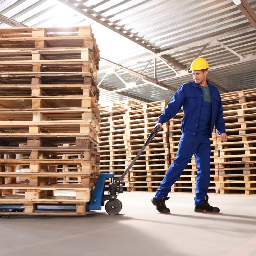 A man is pushing a hand truck in a warehouse filled with wooden pallets.
