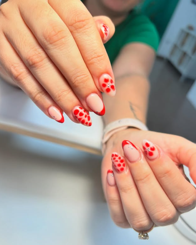 Manos con uñas de forma ovalada que presentan una punta francesa roja y un patrón de pequeños puntos rojos sobre un fondo nude.