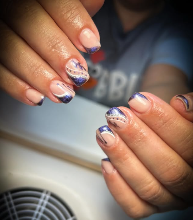 Primer plano de manos con uñas cuidadas. Las uñas presentan una base transparente con puntas moradas y diseños acentuados con destellos y líneas.