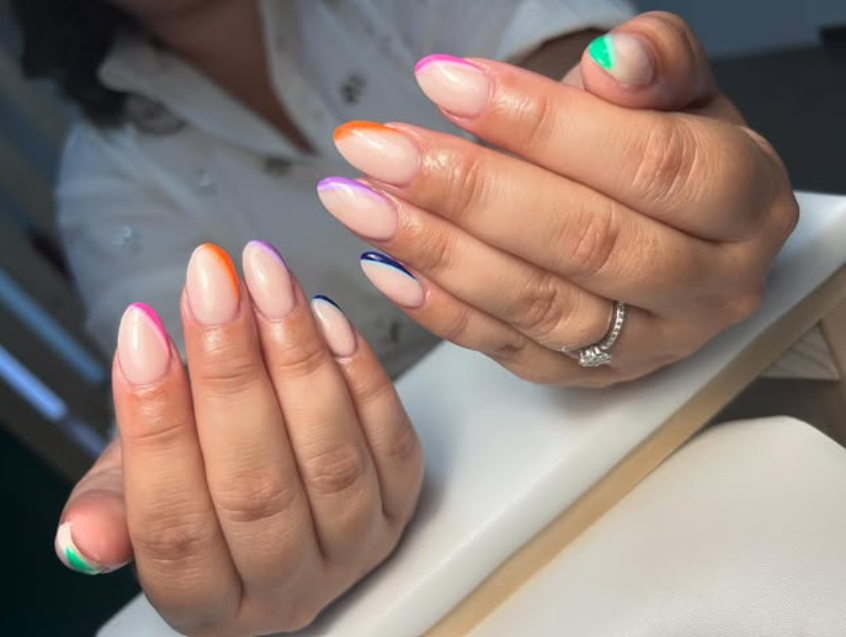 Manos con uñas ovaladas, cada una con un borde de un color brillante diferente. Una mano lleva un anillo con un diamante.