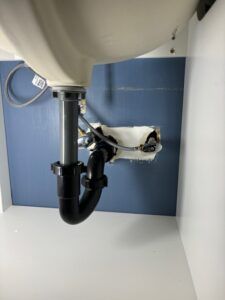 Under-sink view showing a black P-trap connected to a drain pipe and water supply lines against a blue wall.