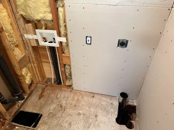 Unfinished laundry room wall with plumbing connections, electrical outlet, and floor drains exposed in wood-framed structure.