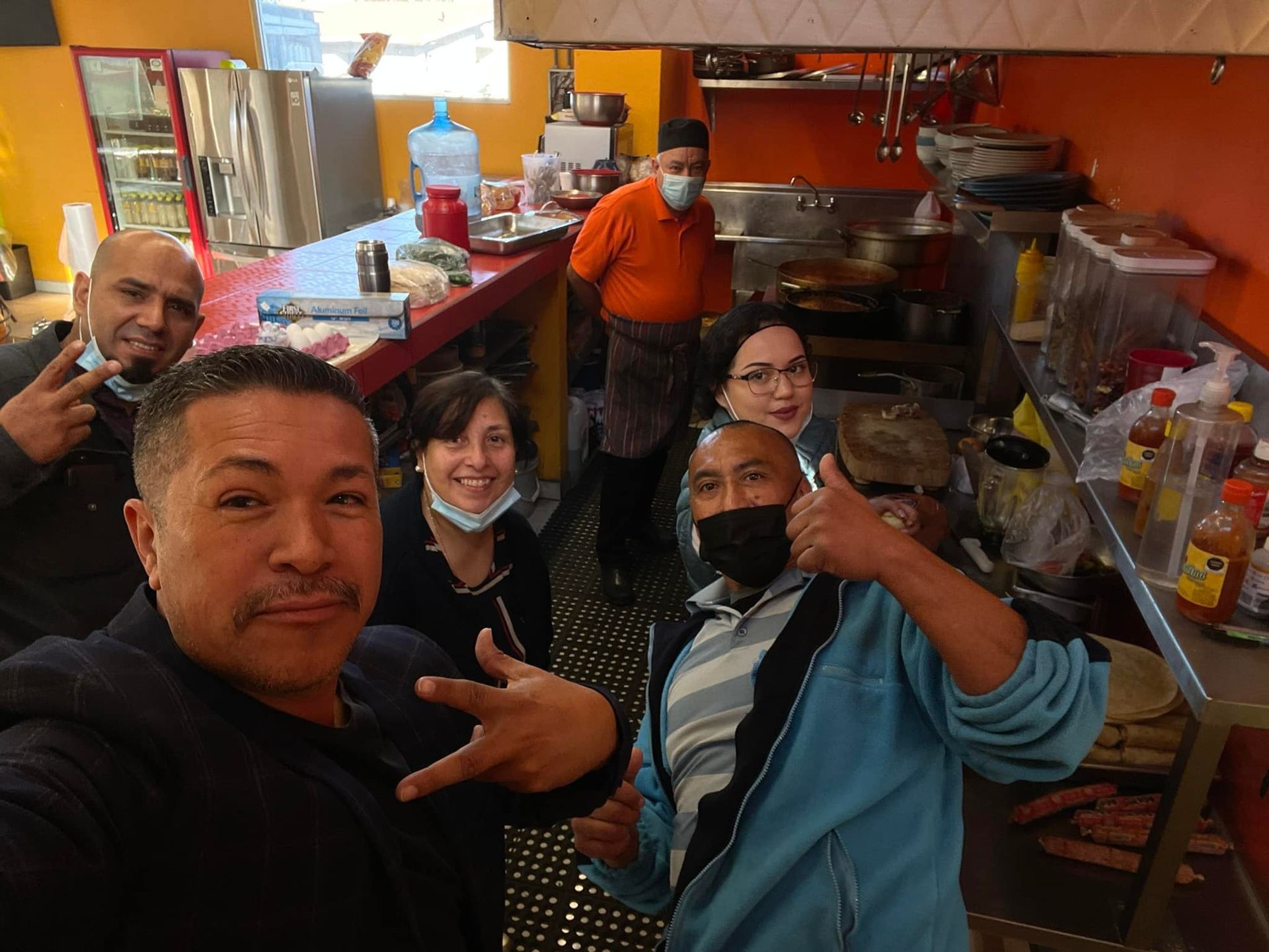 A group of five people poses for a selfie inside a kitchen, some smiling or gesturing, with stainless steel equipment.