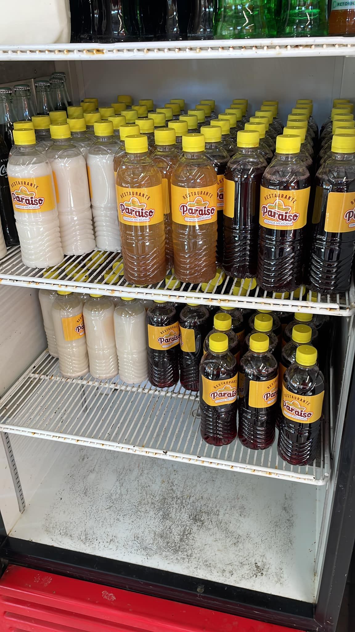 Bottles of various colored drinks with yellow caps are arranged on two shelves inside a commercial refrigerator.