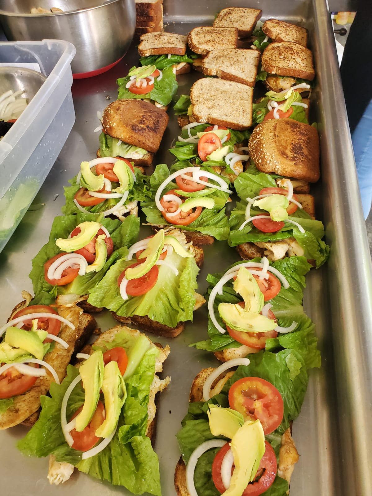 Open-faced sandwiches with lettuce, tomato, onion, and avocado slices arranged in rows on a metal catering tray.