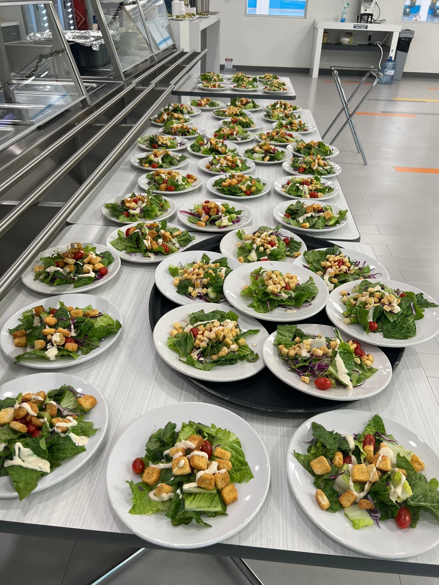 Many white plates topped with green salads, croutons, and cherry tomatoes are arranged on tables in a cafeteria.