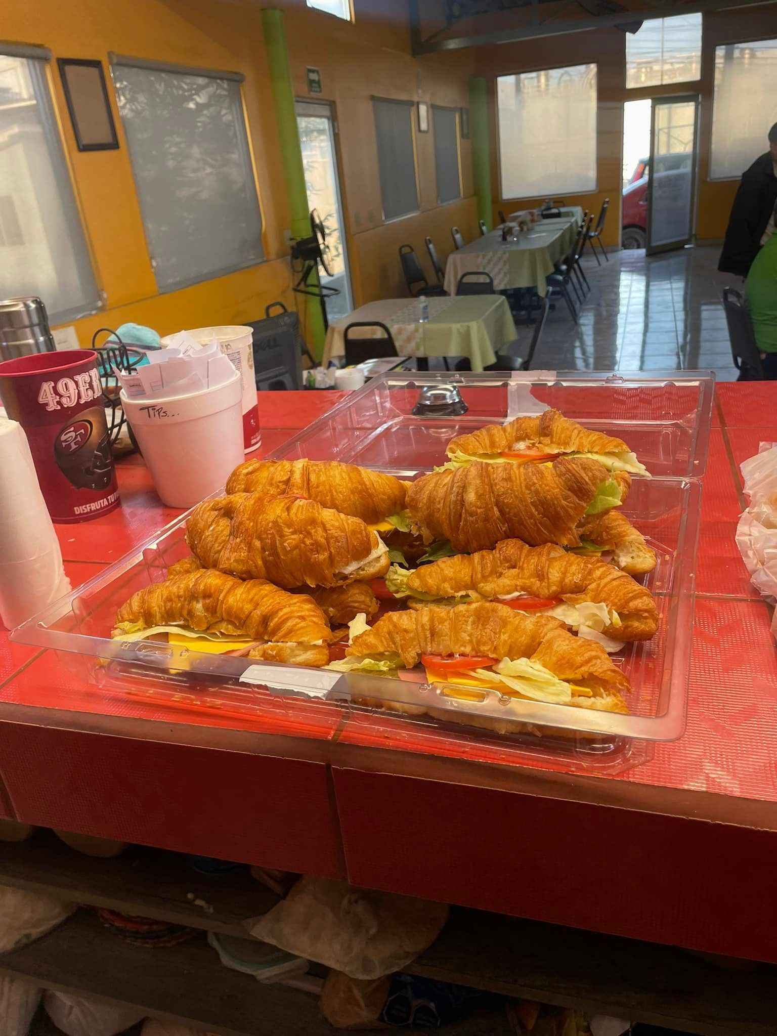 A clear plastic tray holds several croissant sandwiches on a red surface inside a restaurant with tables in the background.