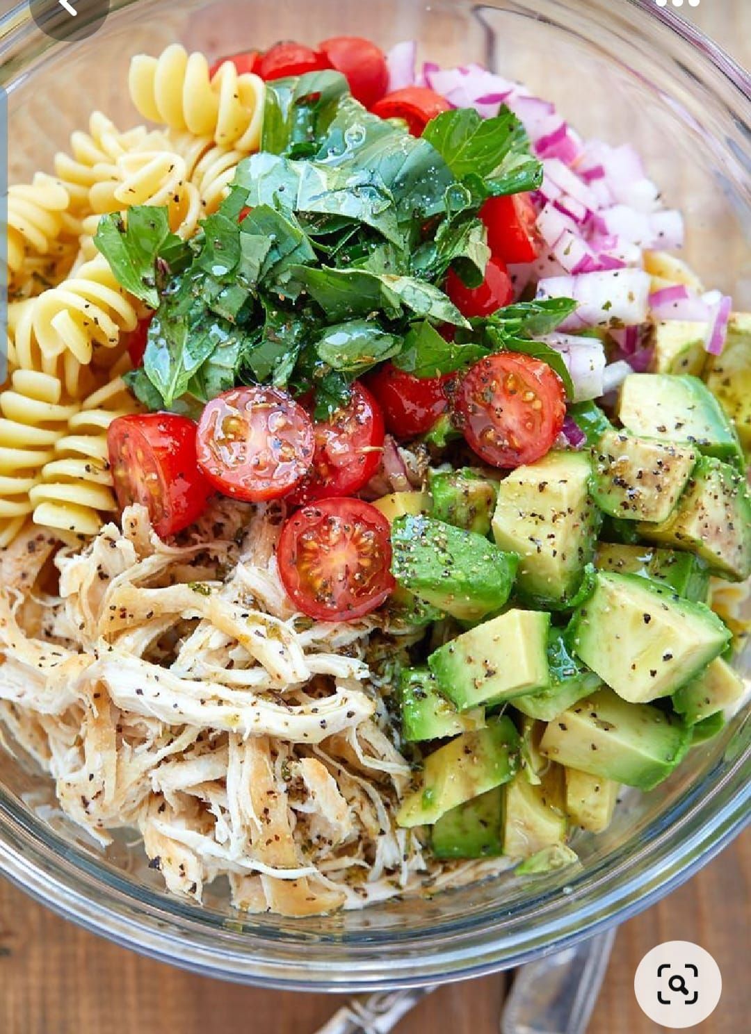 A bowl of pasta salad with shredded chicken, diced avocado, halved cherry tomatoes, red onion, and chopped fresh herbs.