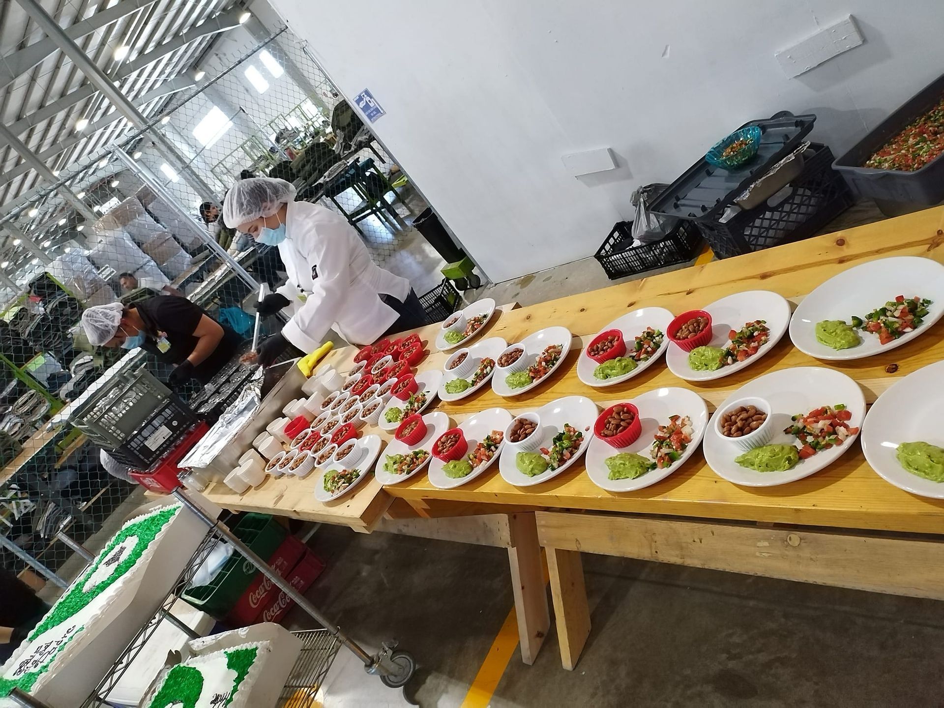 Two workers in hairnets and white uniforms assemble plates of food in a large, industrial-style kitchen.