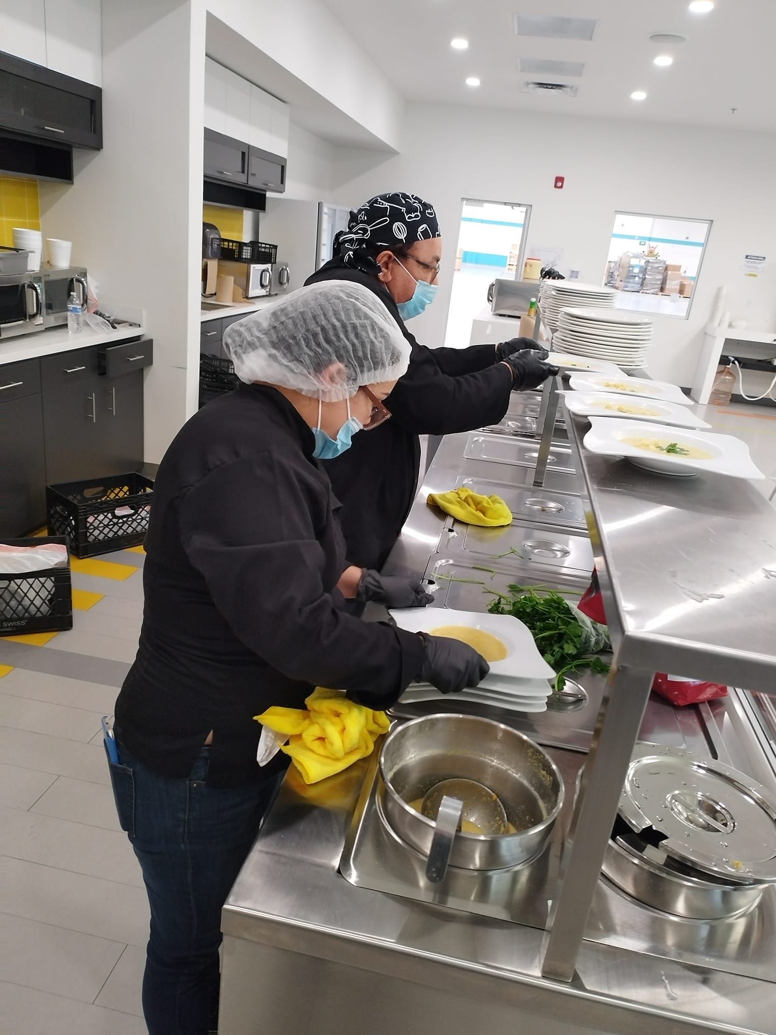 Two kitchen staff in masks and hairnets prepare plates on a long stainless steel service counter in a commercial kitchen.