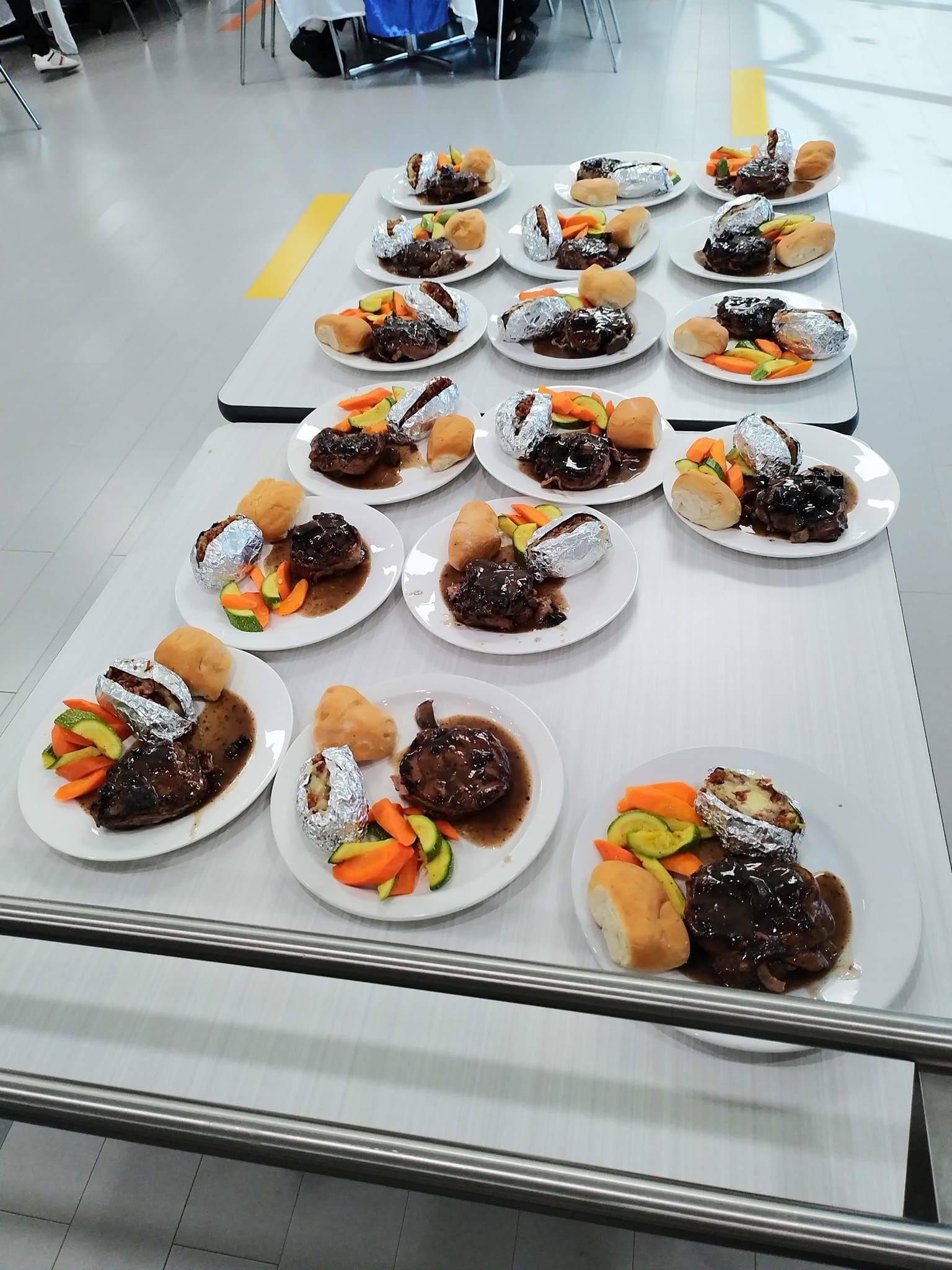 Many plates of steak, potatoes wrapped in foil, and vegetables are arranged on a white table in a dining hall.