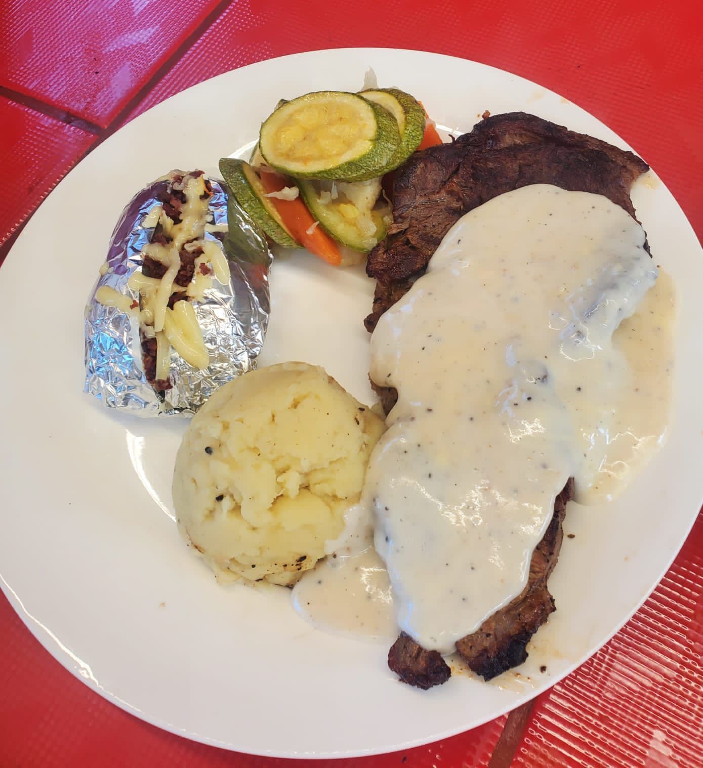 A plate featuring a steak topped with white sauce, mashed potatoes, a foil-wrapped baked potato, and sautéed vegetables.