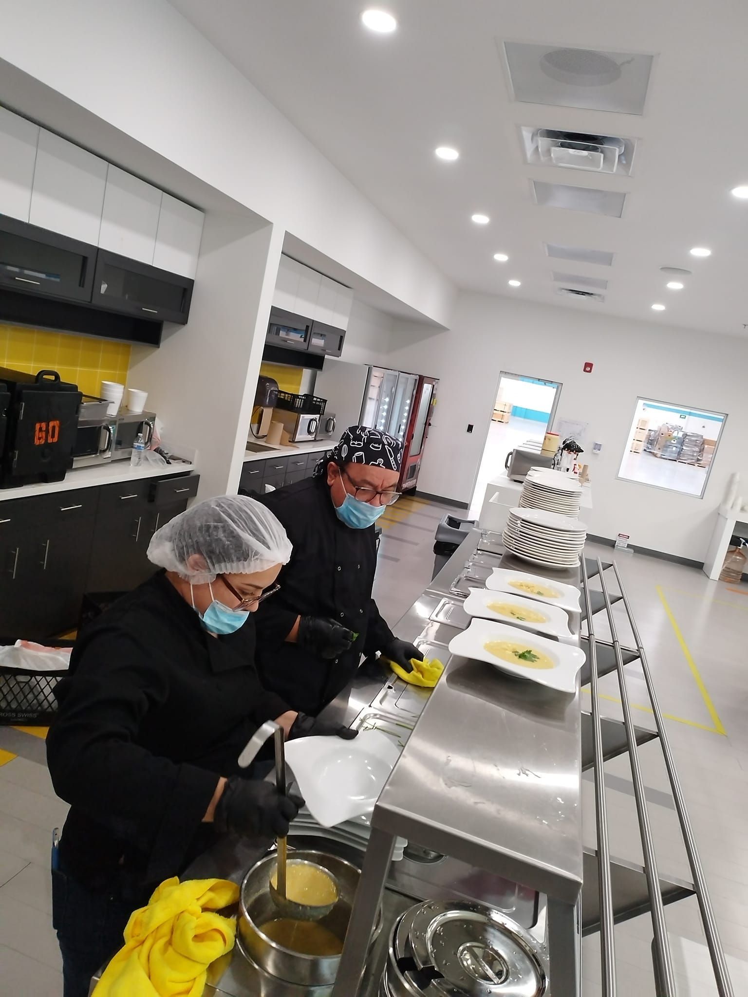 Two kitchen staff in black uniforms, hairnets, and face masks ladle soup into bowls along a stainless steel counter.