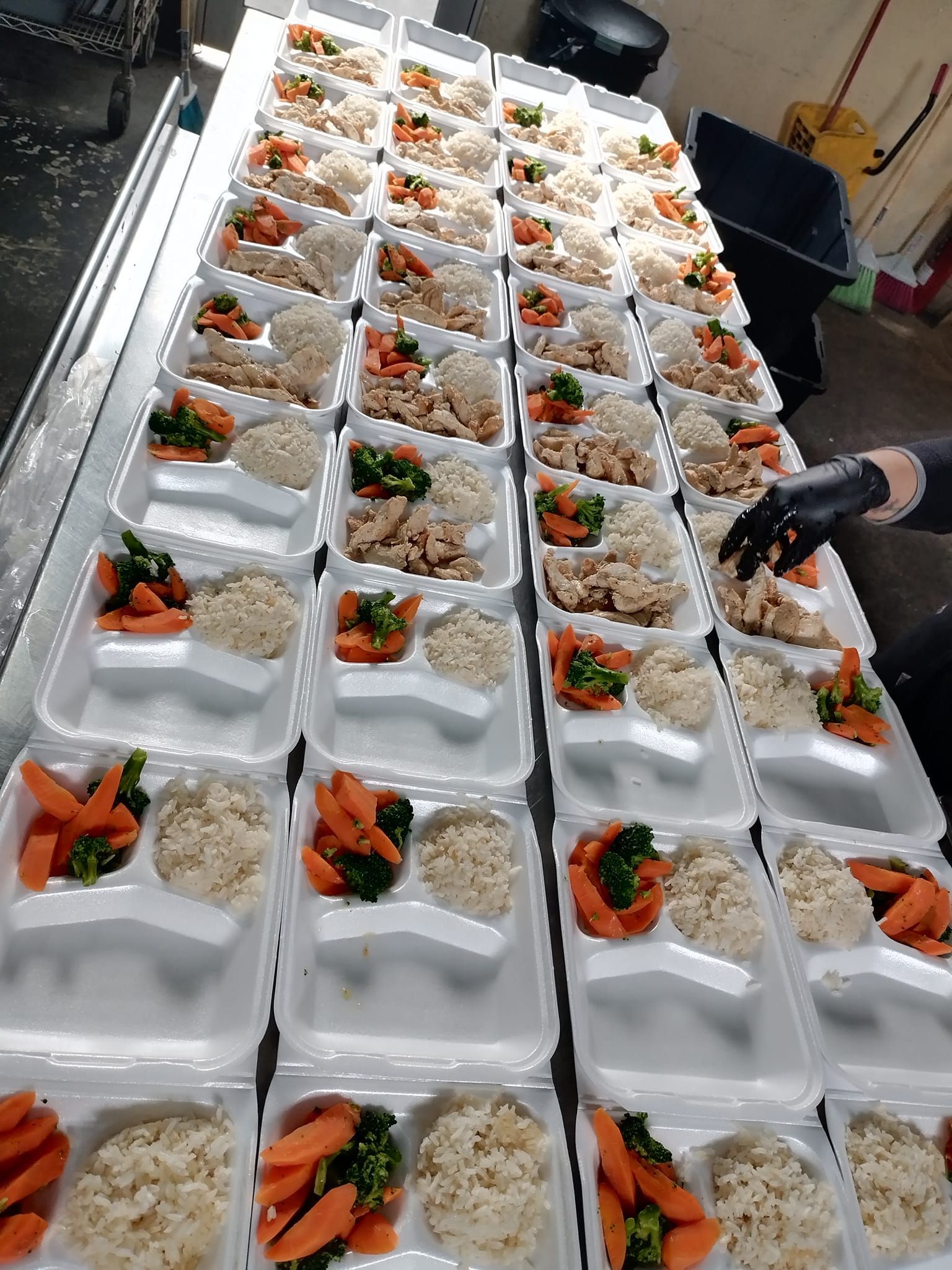 Rows of white styrofoam containers filled with portions of rice and cooked vegetables are arranged on a prep table.