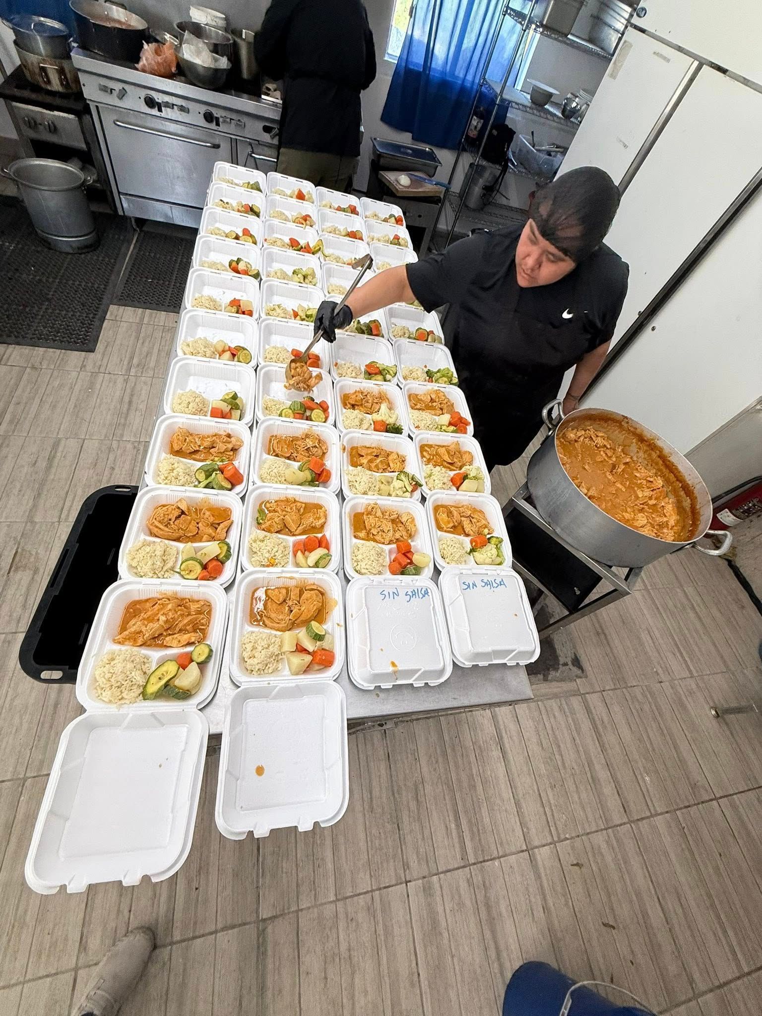 A person in a kitchen fills white takeout containers with food, arranged in long rows on a table next to a large pot.