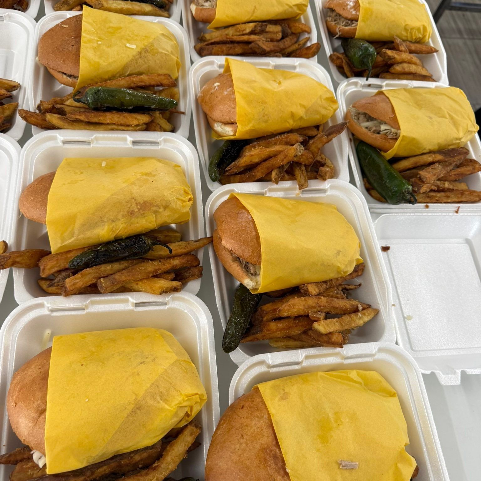 Multiple take-out containers, each holding a burger wrapped in yellow paper, a serving of fries, and a whole jalapeño.