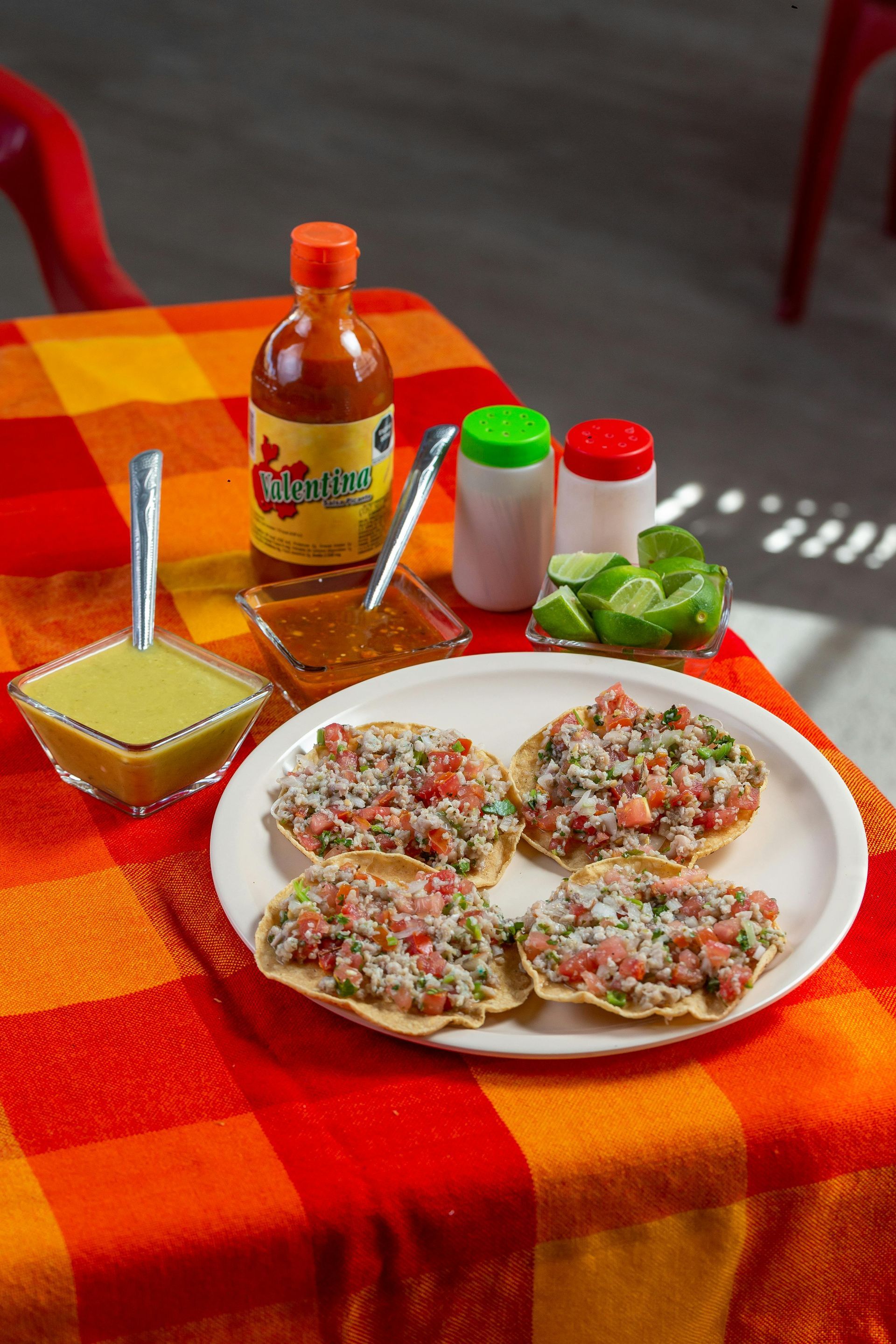 Four tostadas topped with ceviche on a white plate, served with lime wedges, two sauces, and seasonings on a plaid table.