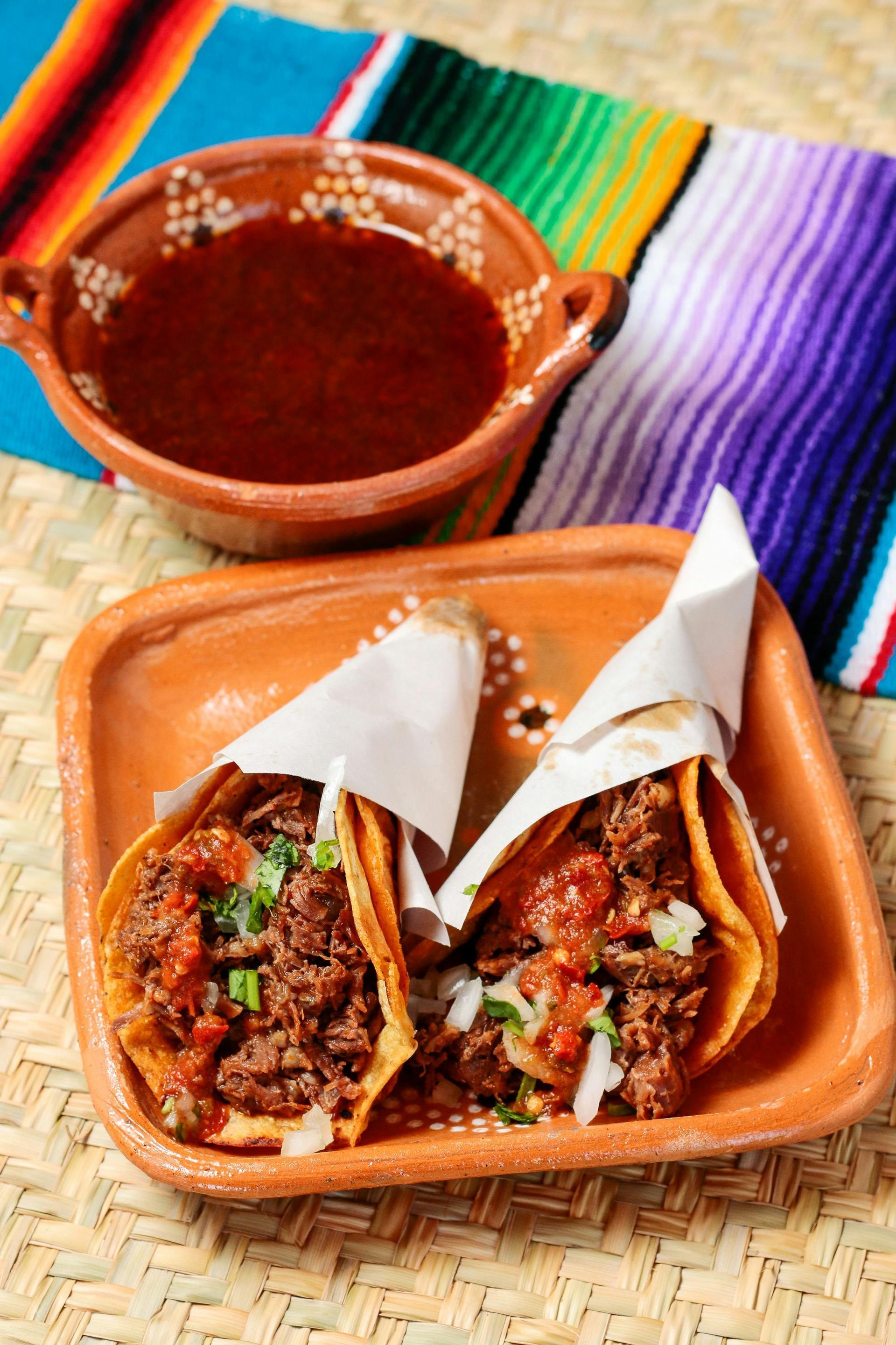 Two tacos in paper wrappers and a bowl of dipping sauce on a plate atop a woven mat with a colorful striped cloth.