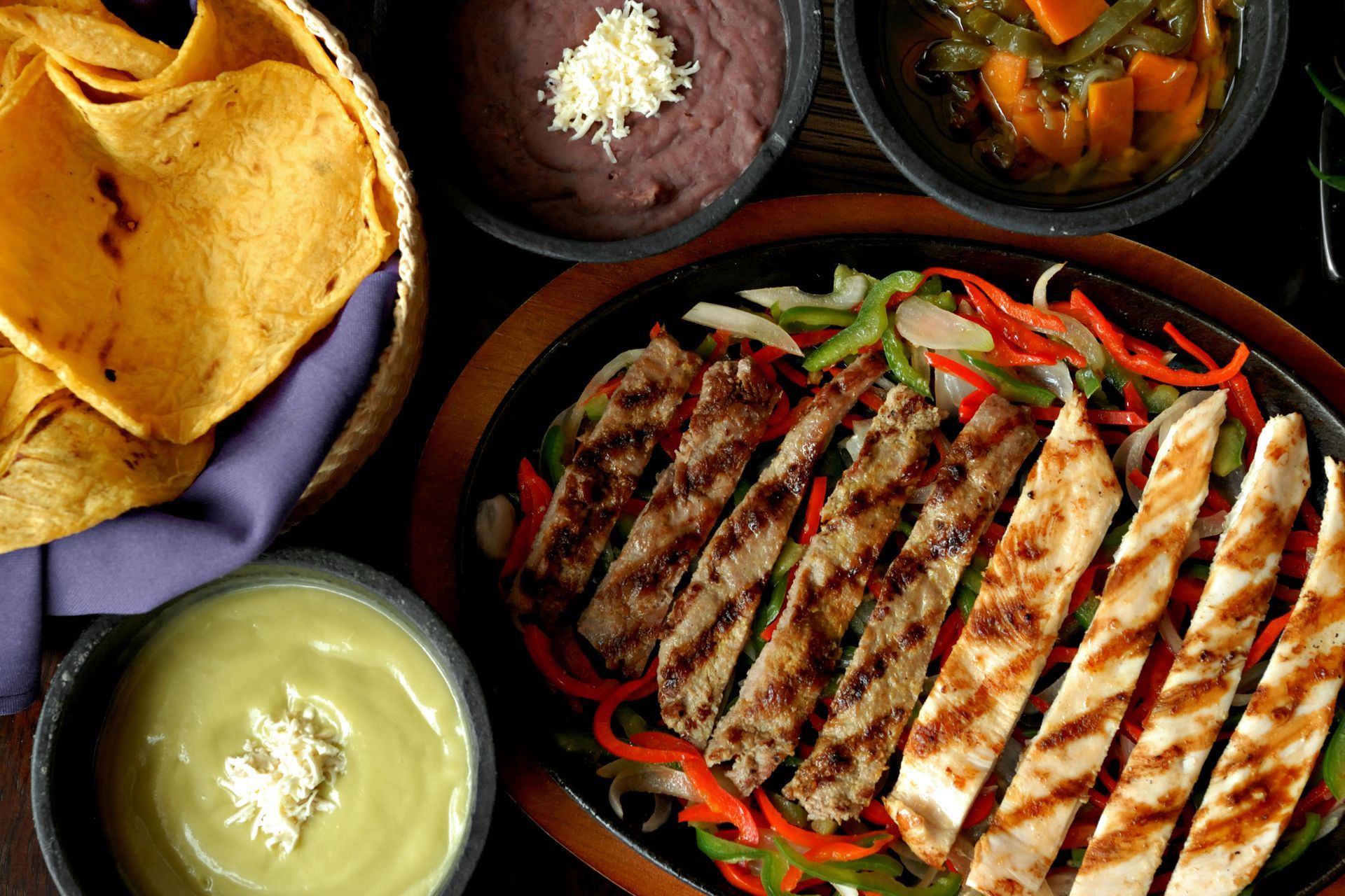 A platter of grilled chicken and beef fajitas with peppers, onions, tortillas, beans, guacamole, and salsa.
