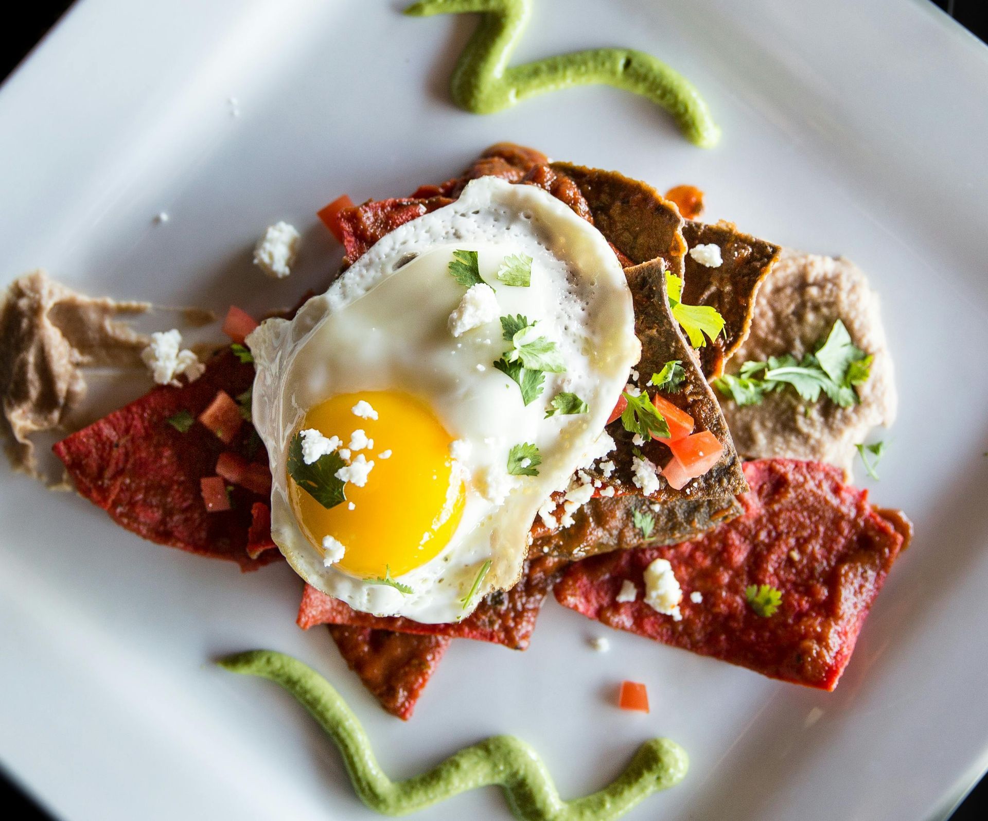 A sunny-side-up egg over colorful tortilla chips, beans, and fresh herbs, garnished with zig-zags of green sauce.