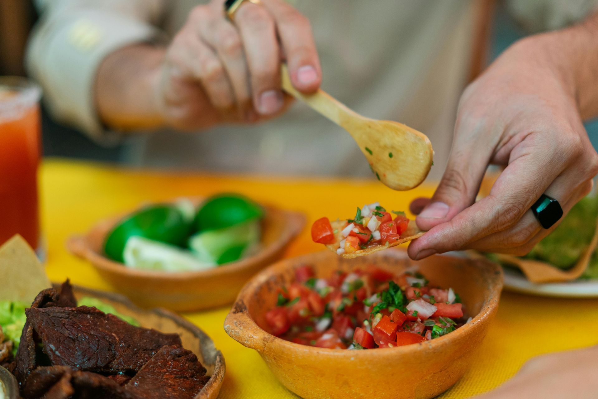 A person uses a wooden spoon to place fresh salsa onto a tortilla chip over a yellow table filled with Mexican food.