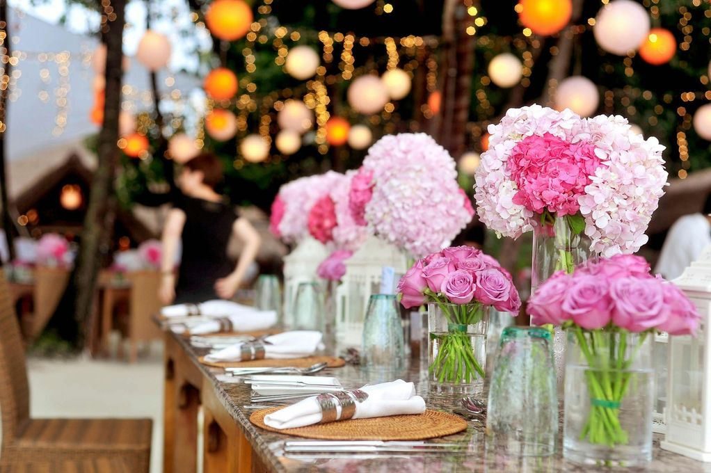 A table set for a celebration with pink floral arrangements, set outdoors with hanging lanterns.