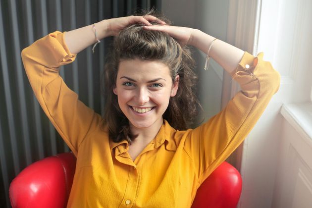 Woman in yellow shirt smiling, arms raised, touching her hair, sitting in a red chair.