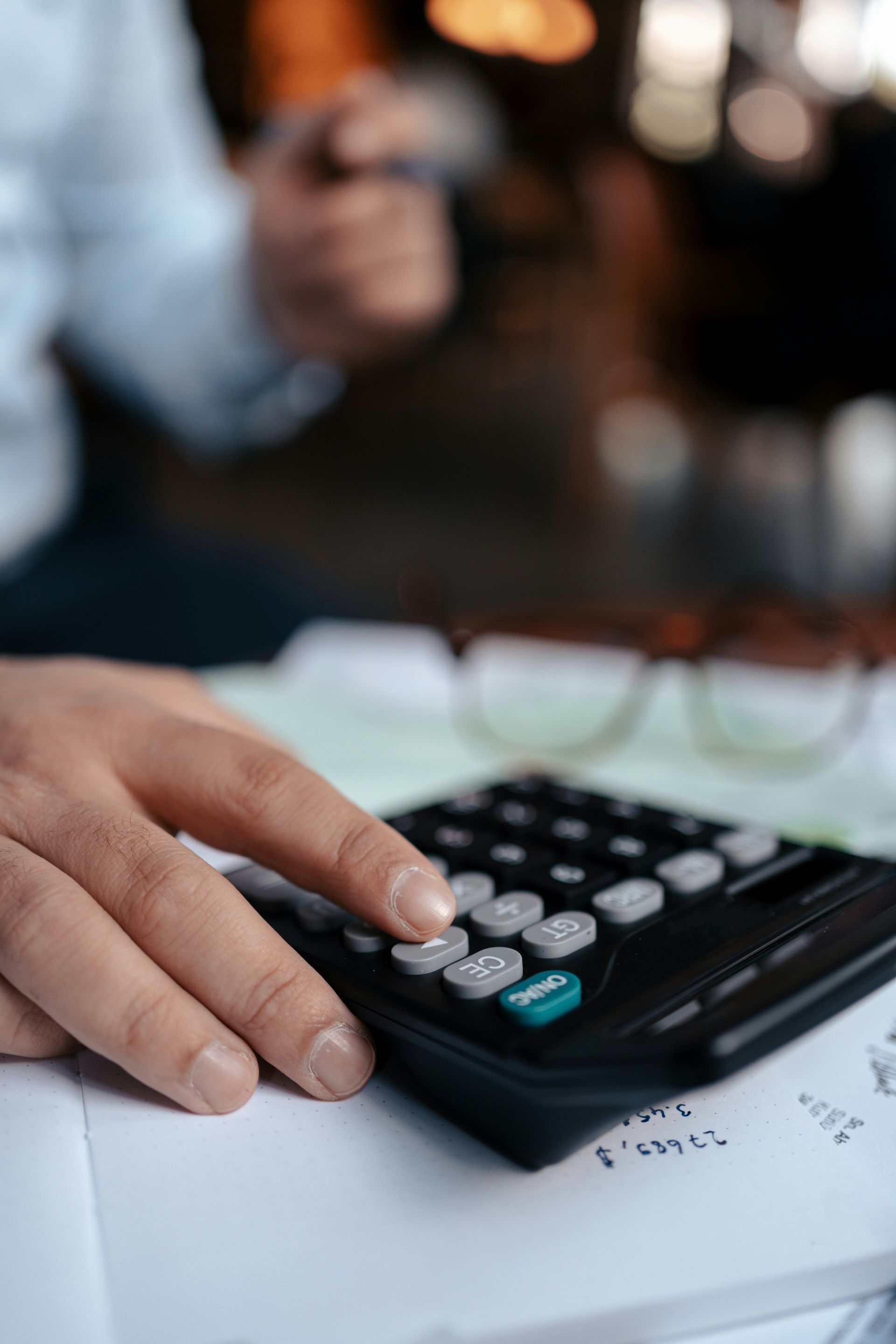 Person's hand using a calculator on a desk with paperwork and glasses in the background.