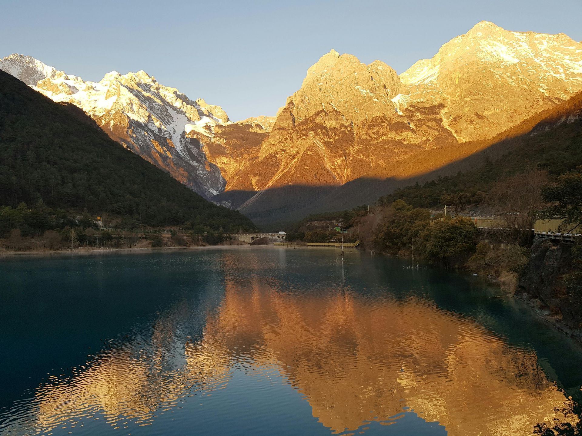 Golden sunlight bathes snow-capped mountains and reflects on calm turquoise lake.