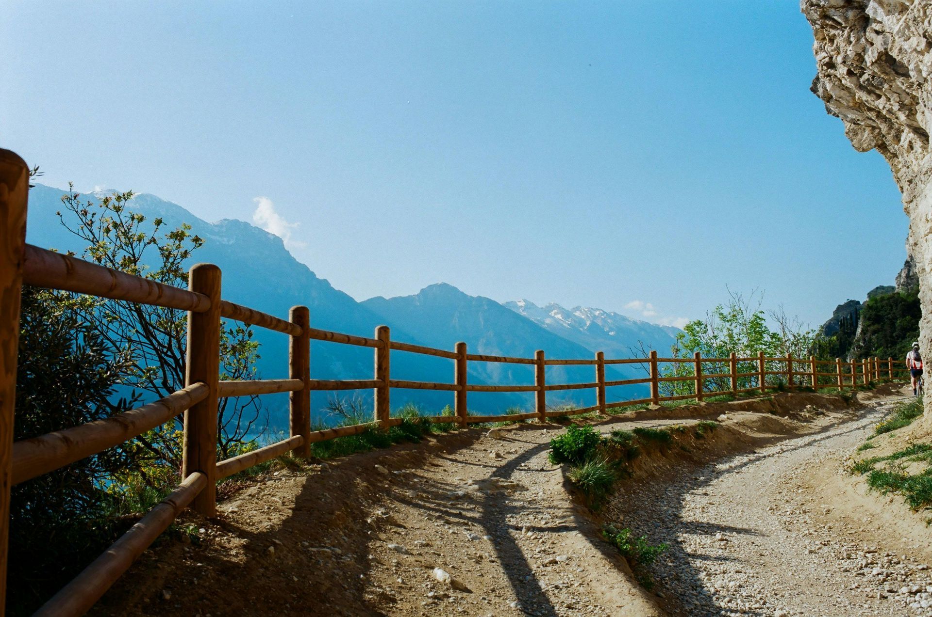 Trail with wooden fence, alongside a cliff with mountain backdrop.