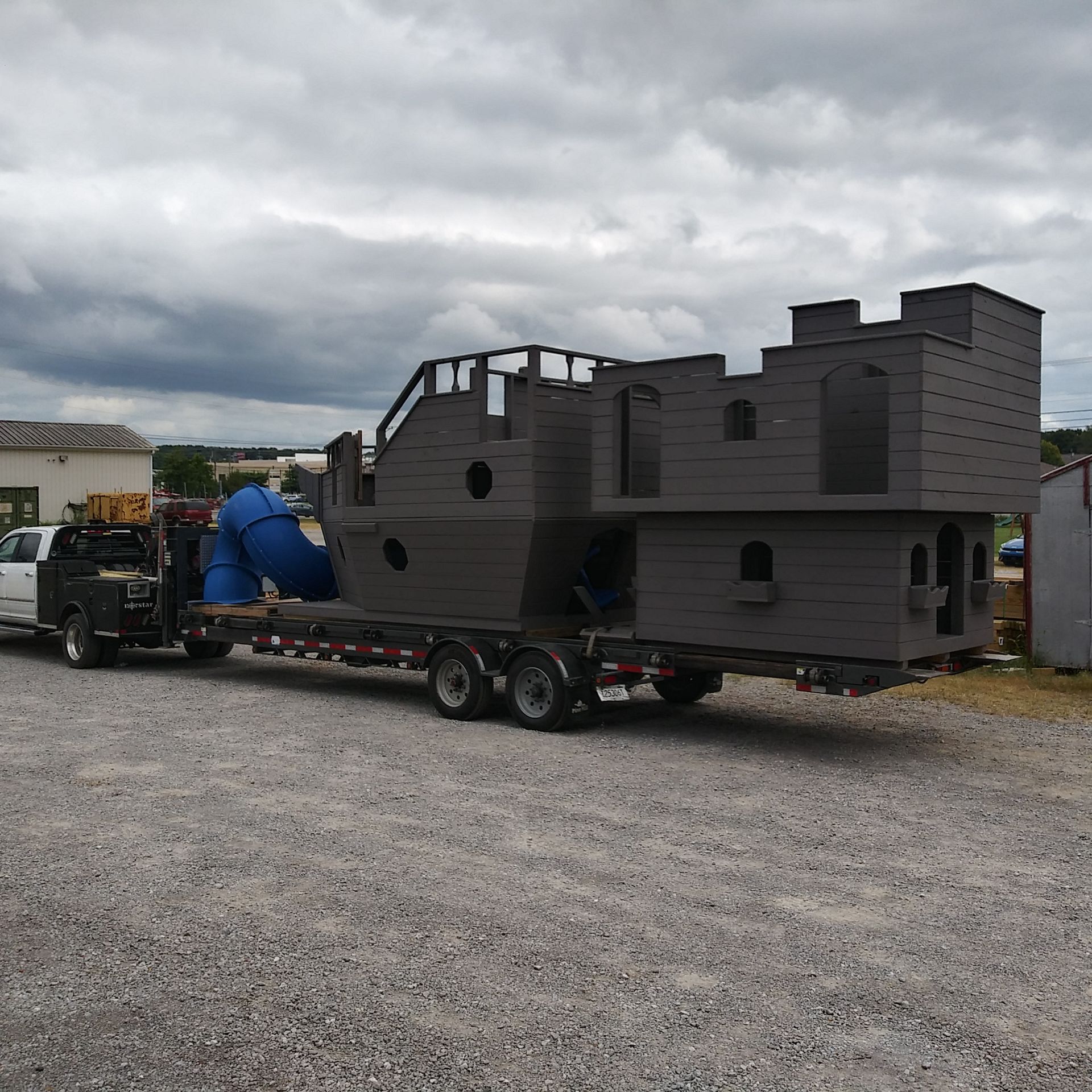 Large gray castle-shaped playset loaded on a trailer, being towed by a truck on gravel.