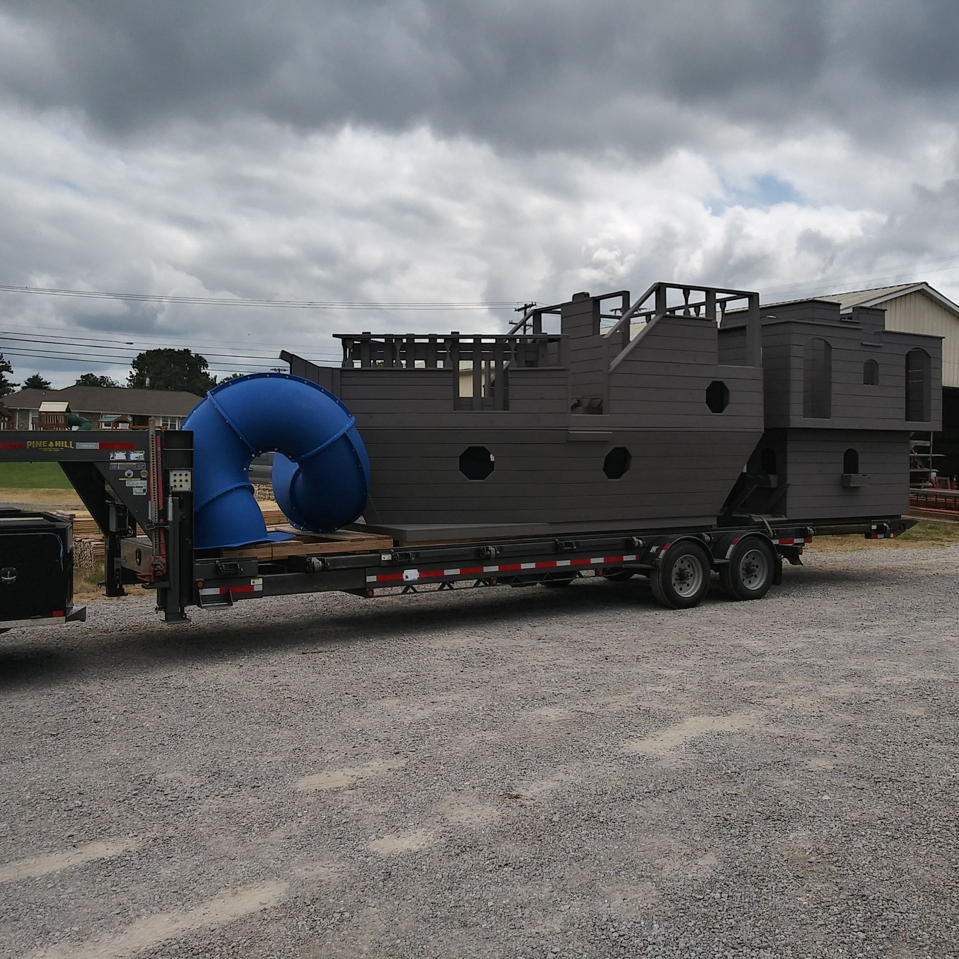 Large wooden pirate ship playground on a trailer, with a blue slide, under a cloudy sky.