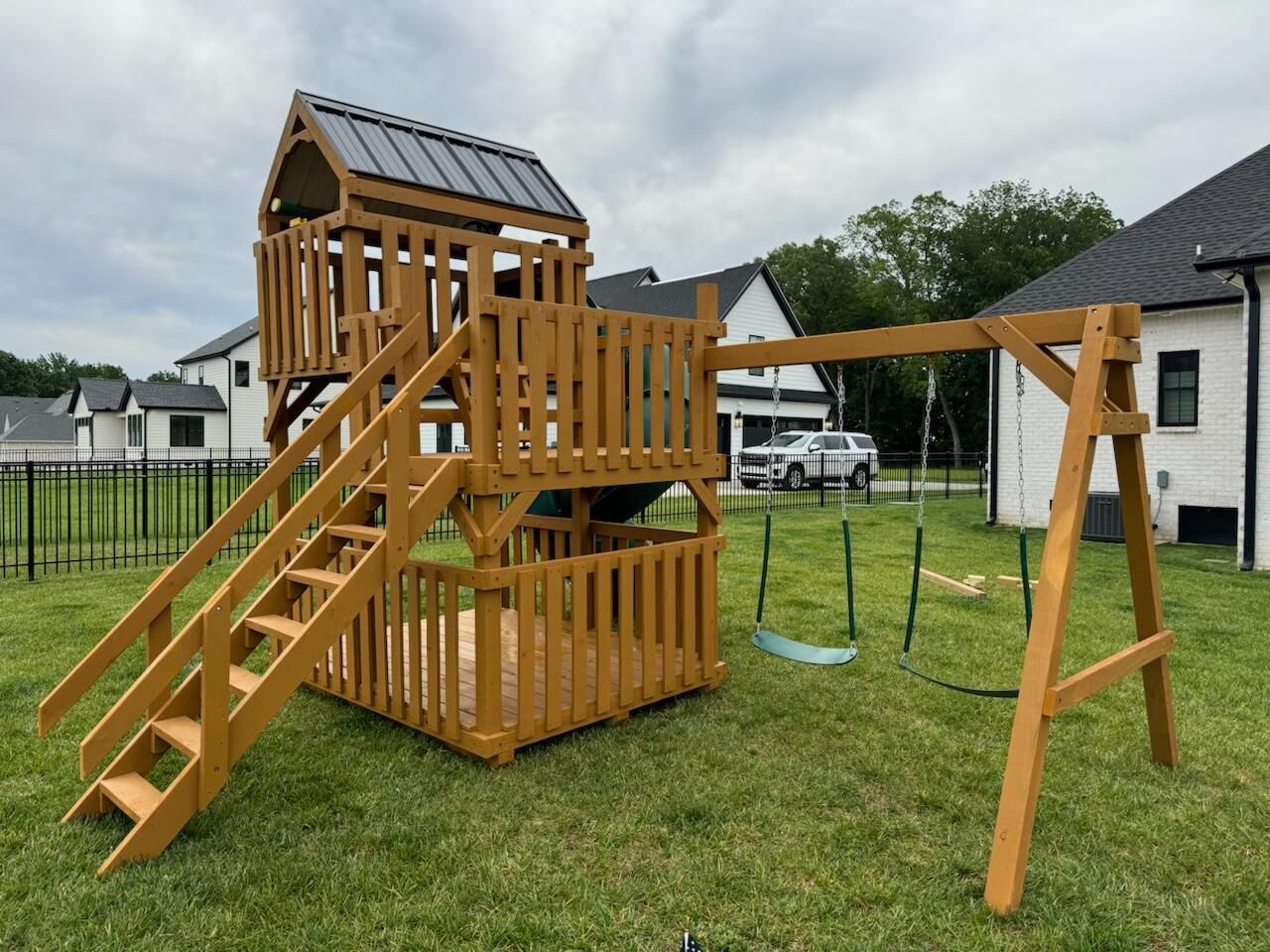 Wooden backyard playset with stairs, slide, and swing set on a grassy lawn under cloudy skies