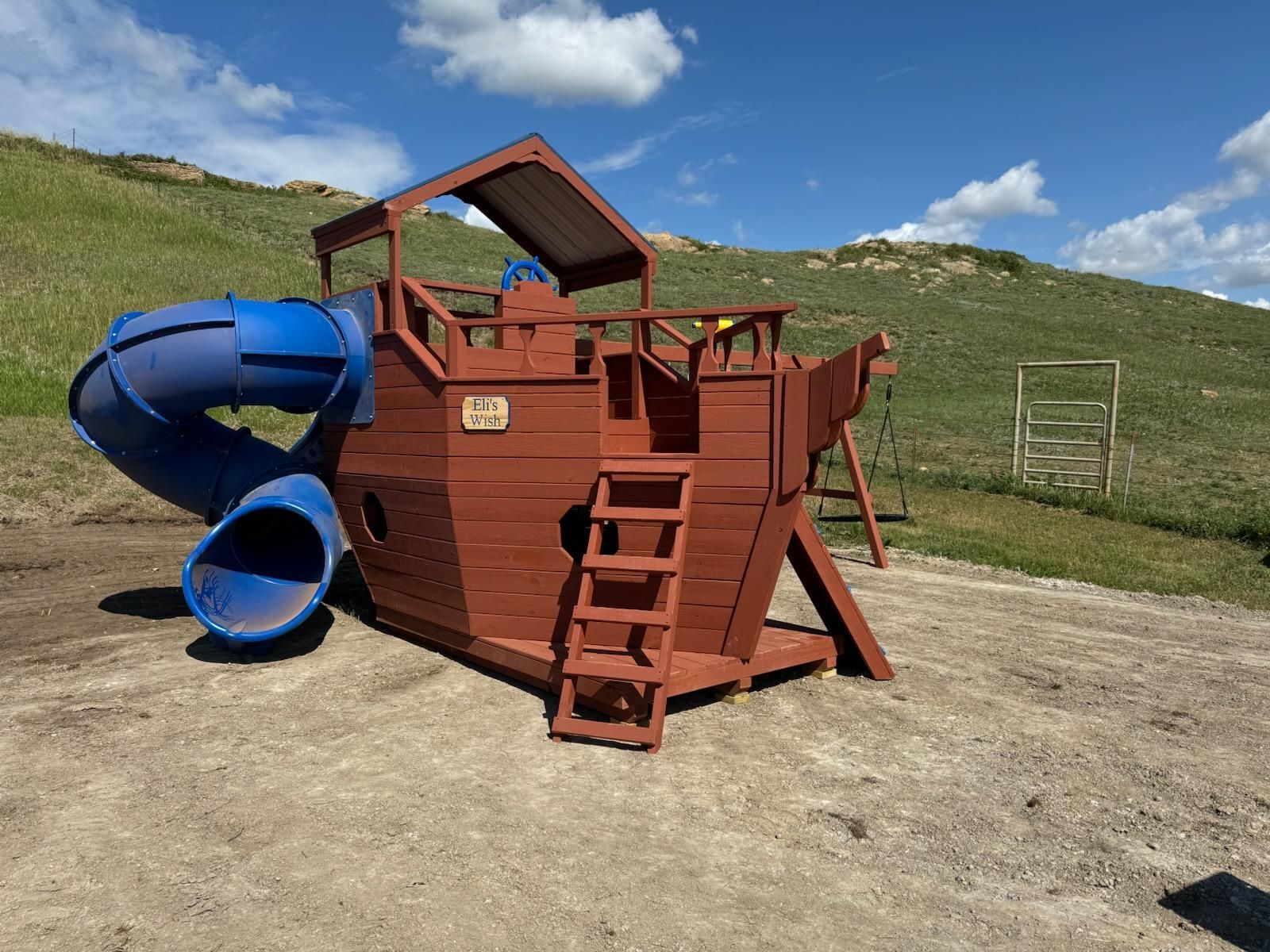 Blue and red playground fort with slides on a dirt lot in a grassy field under a cloudy sky