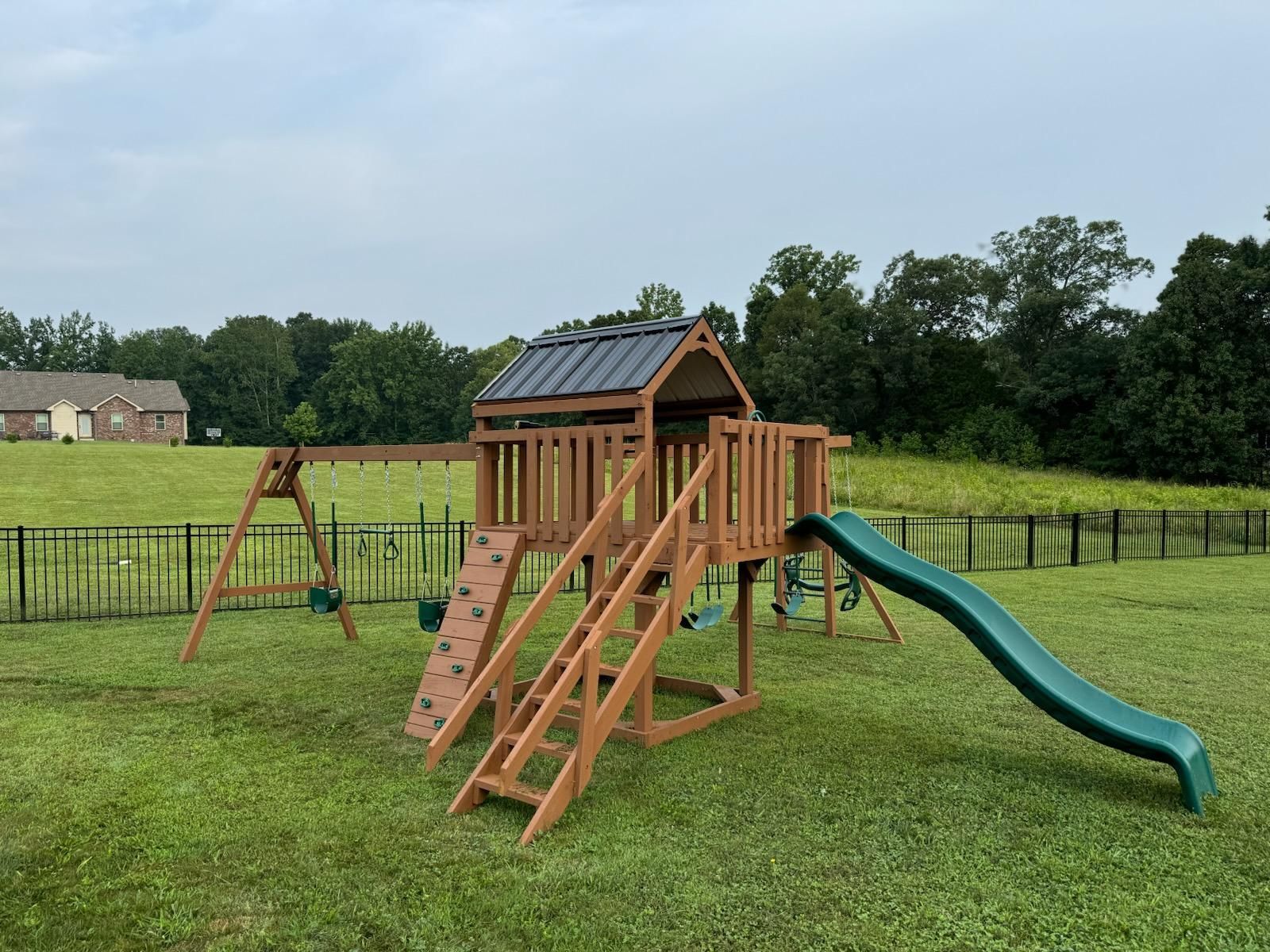 A wooden backyard playset with a green slide, climbing wall, swings, and a roof, set in a grassy field.
