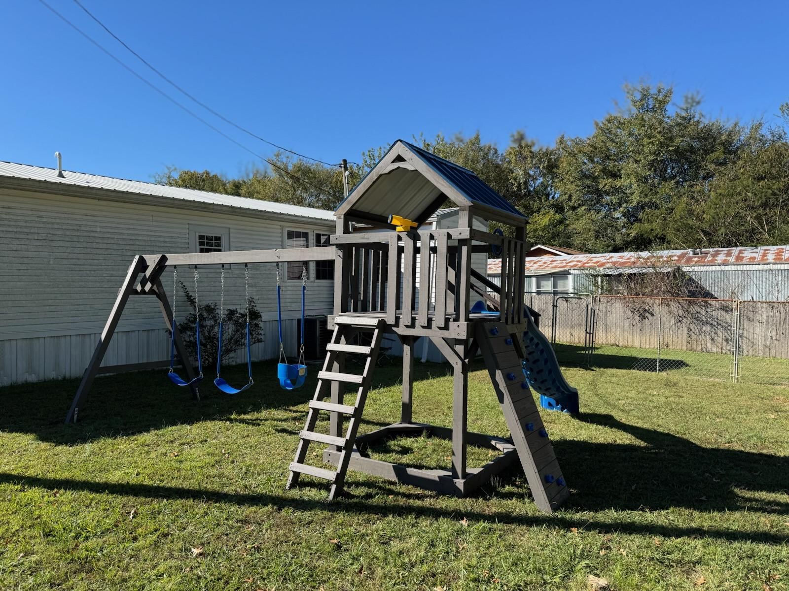 Backyard playset with ladder, slide, and swings on a sunny lawn beside a white house.