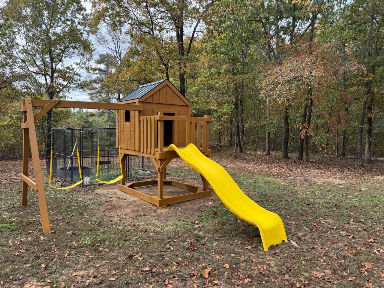Wooden playset with a yellow slide, swings, and a small trampoline in a wooded backyard.