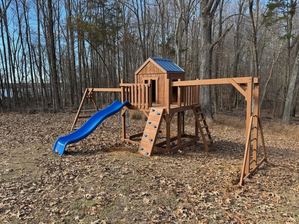 Wooden playset with blue slide, climbing wall, swings, and a treehouse in a wooded area.