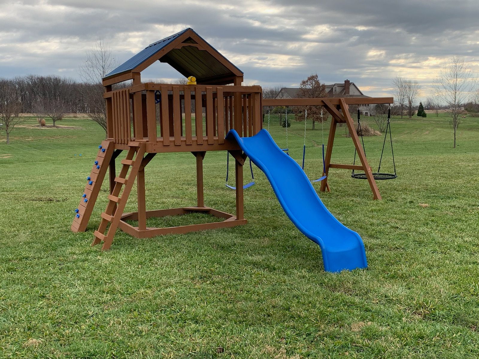 Wooden backyard playset with a blue slide on a grassy field under an overcast sky