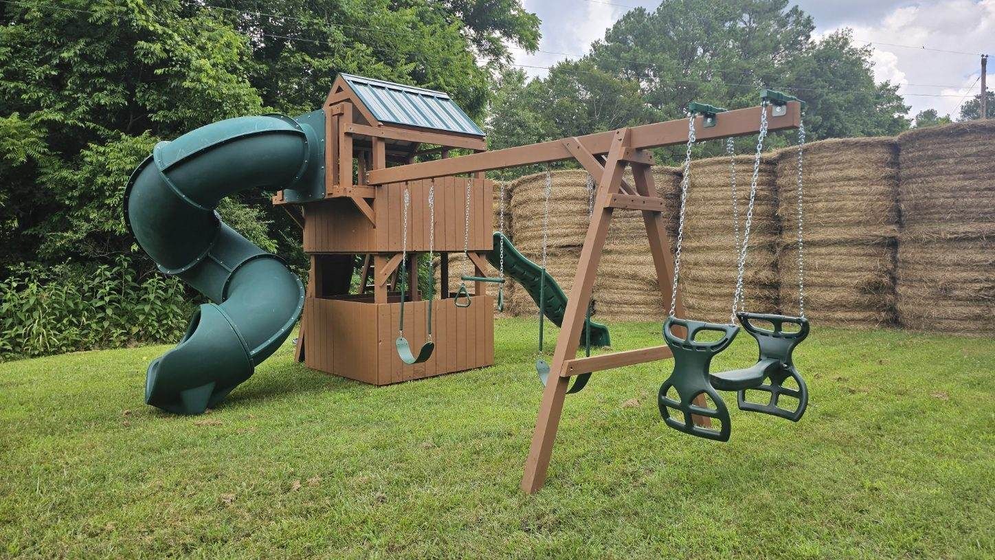 Wooden playground with a green slide, swings, and a playhouse, in a grassy backyard.