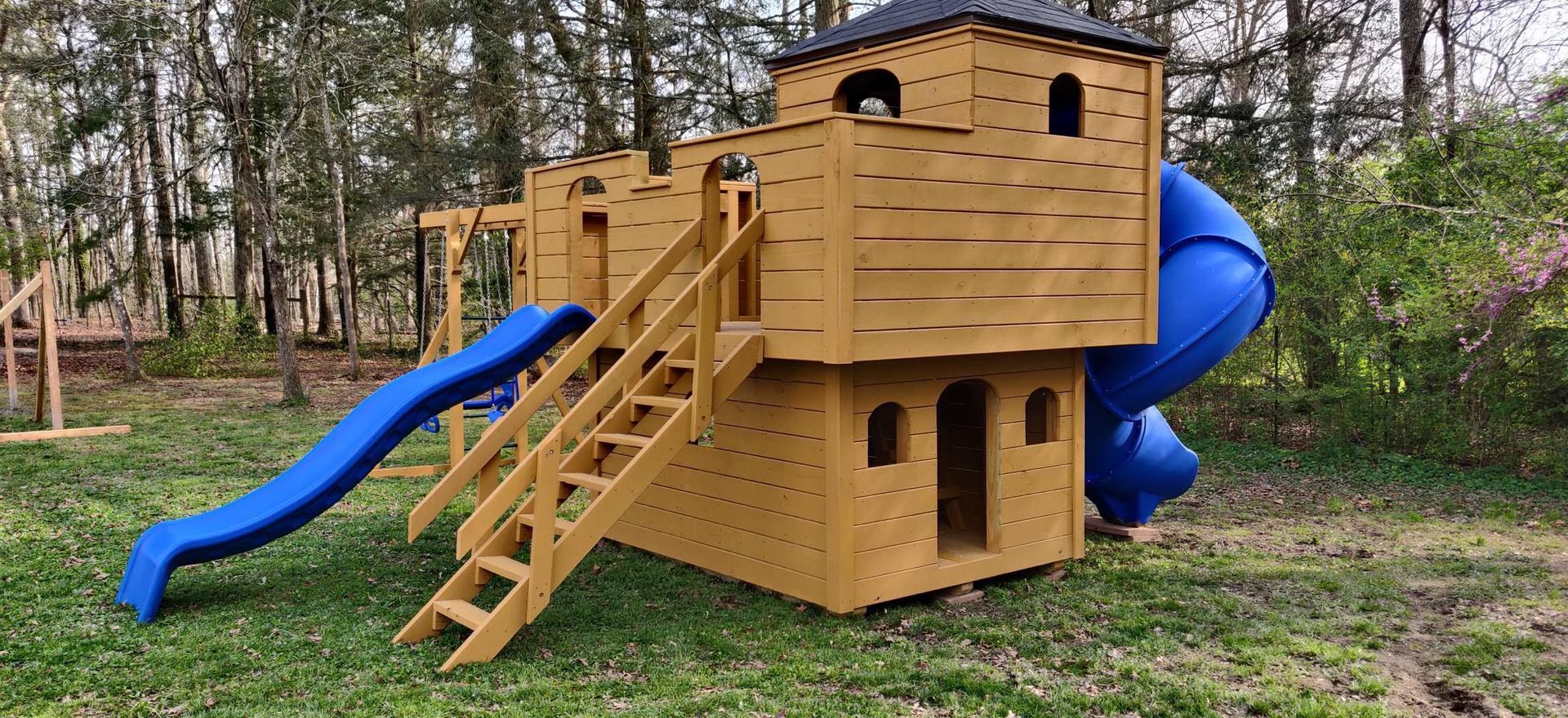 Wooden play structure with blue slides and stairs, set in a grassy yard.