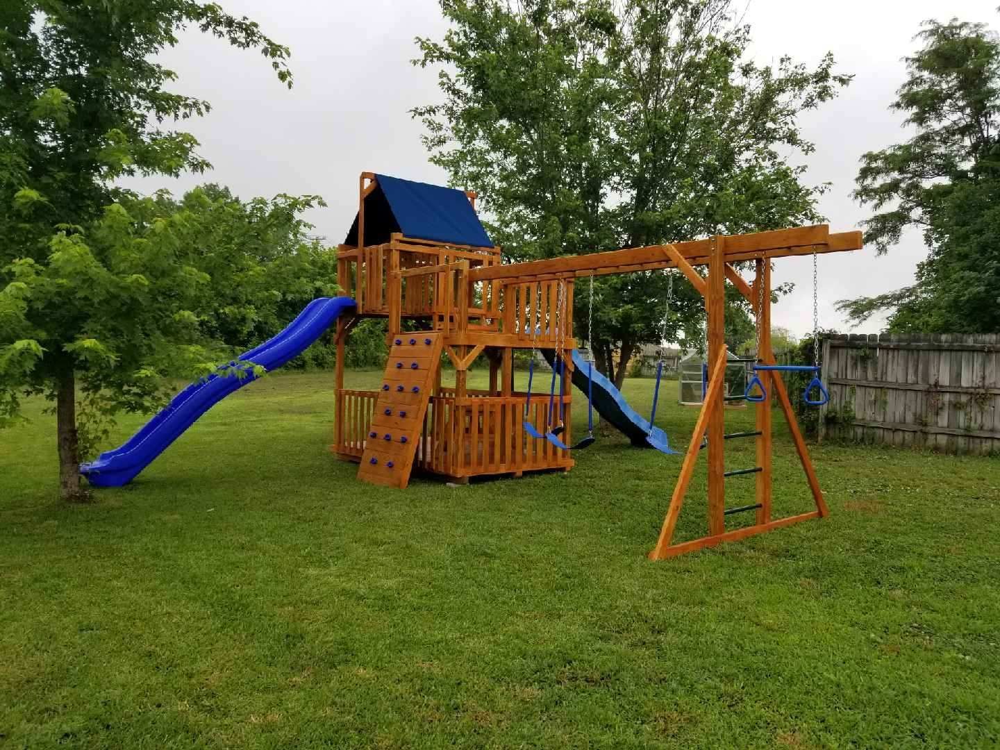 Wooden backyard playground set with a blue slide, rock-climbing wall, swings, and a blue canopy on a grassy lawn.