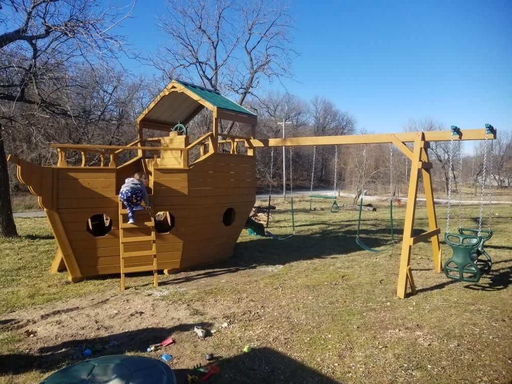 Child climbing a wooden ship-shaped playground structure with a swing set in a park on a sunny day.
