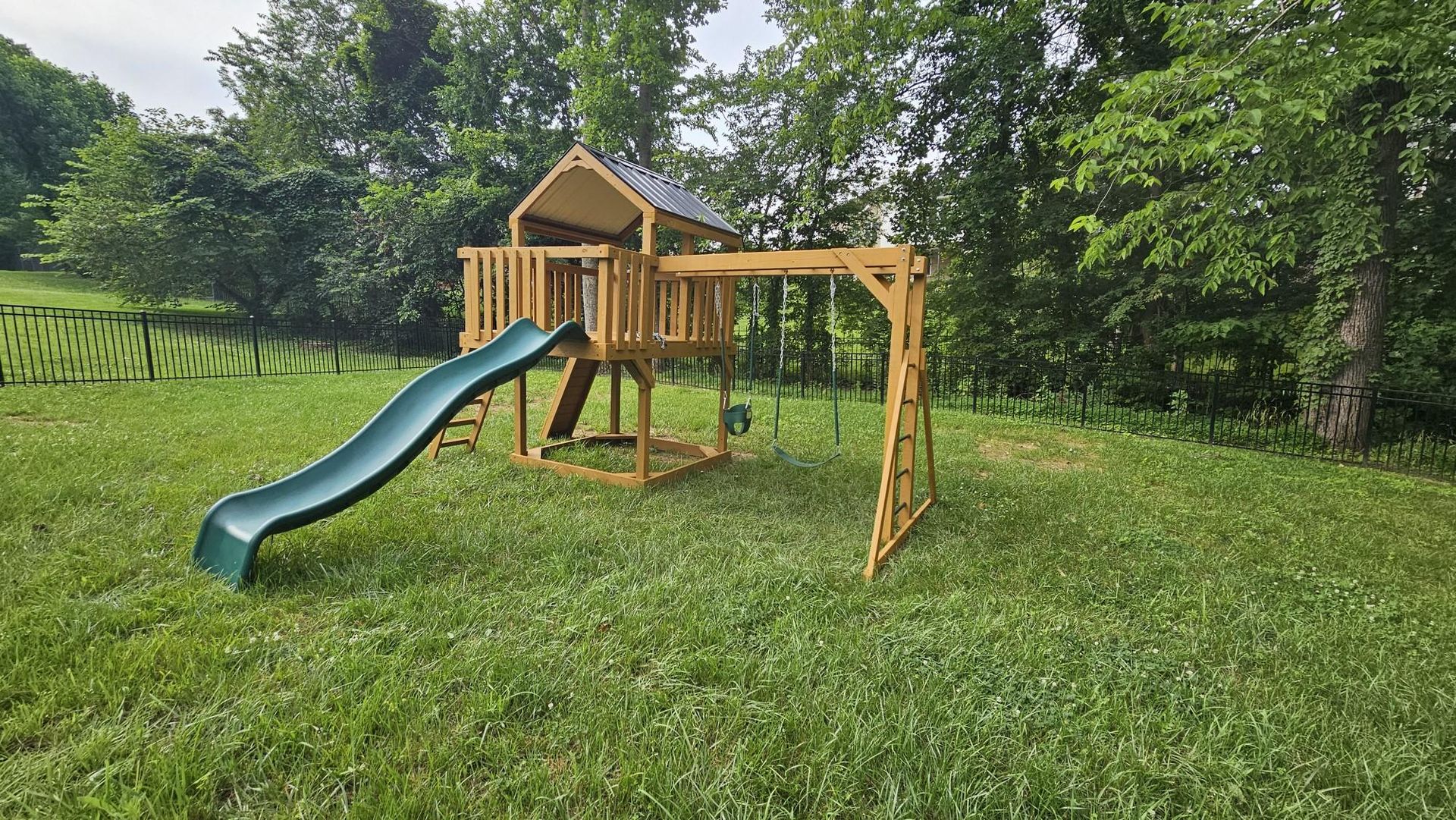 Wooden backyard playset with green slide on a grassy lawn, surrounded by trees.