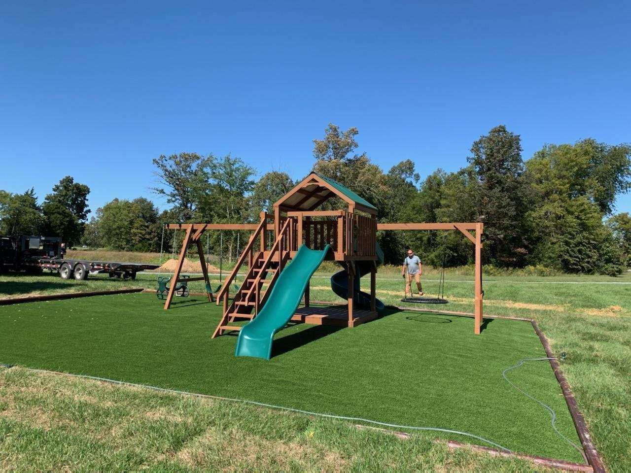 Playground with slide, swings, and playhouse on artificial turf. Man stands nearby on a sunny day.