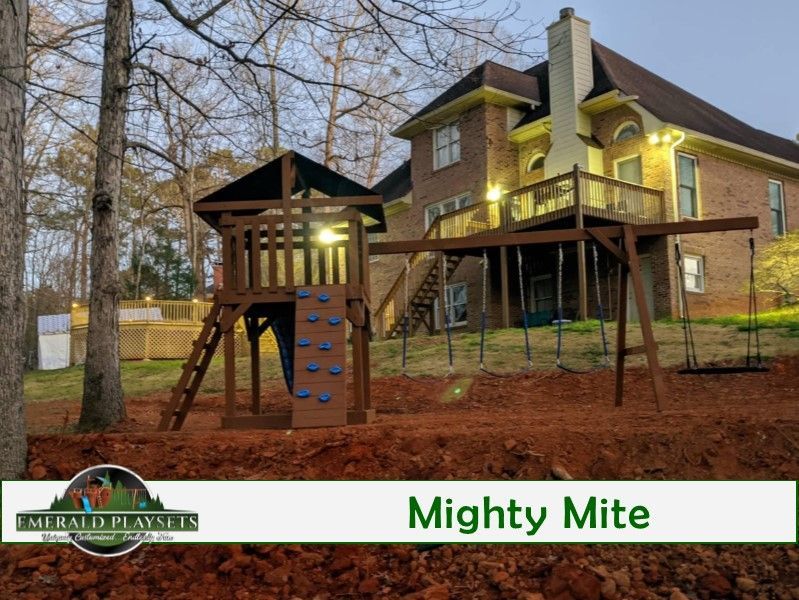 Wooden playground set in a backyard, featuring a playhouse, rock wall, and swingset.