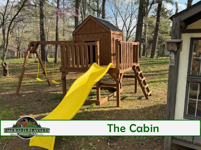 Wooden playset with a cabin, slide, swing set, and a ladder. Set in a grassy yard.