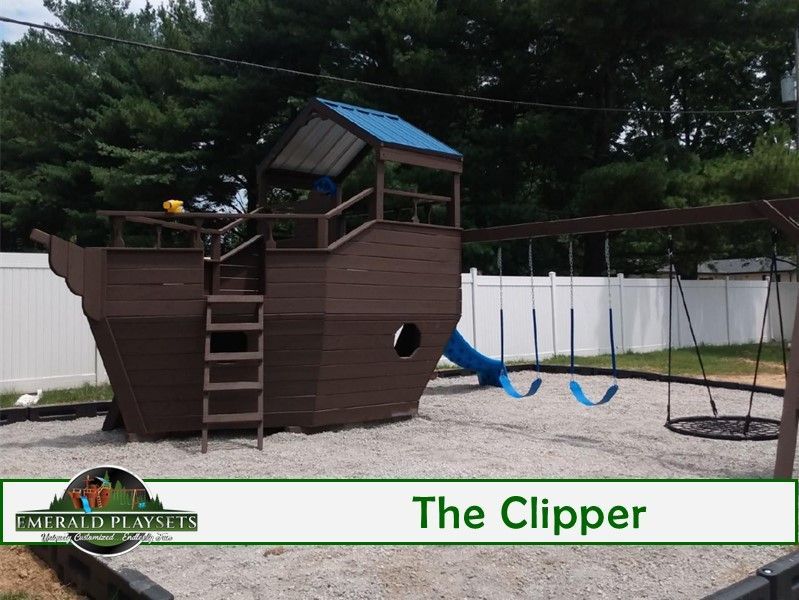 A brown wooden playset shaped like a ship with a blue roof, slide, swings, and tire swing in a yard.